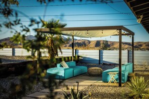 Un área de estar al aire libre con pérgola, cojines y vistas a un lago y montañas.