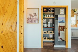 Un pasillo con una puerta de madera, un paisaje desértico enmarcado y un estante con varios objetos.