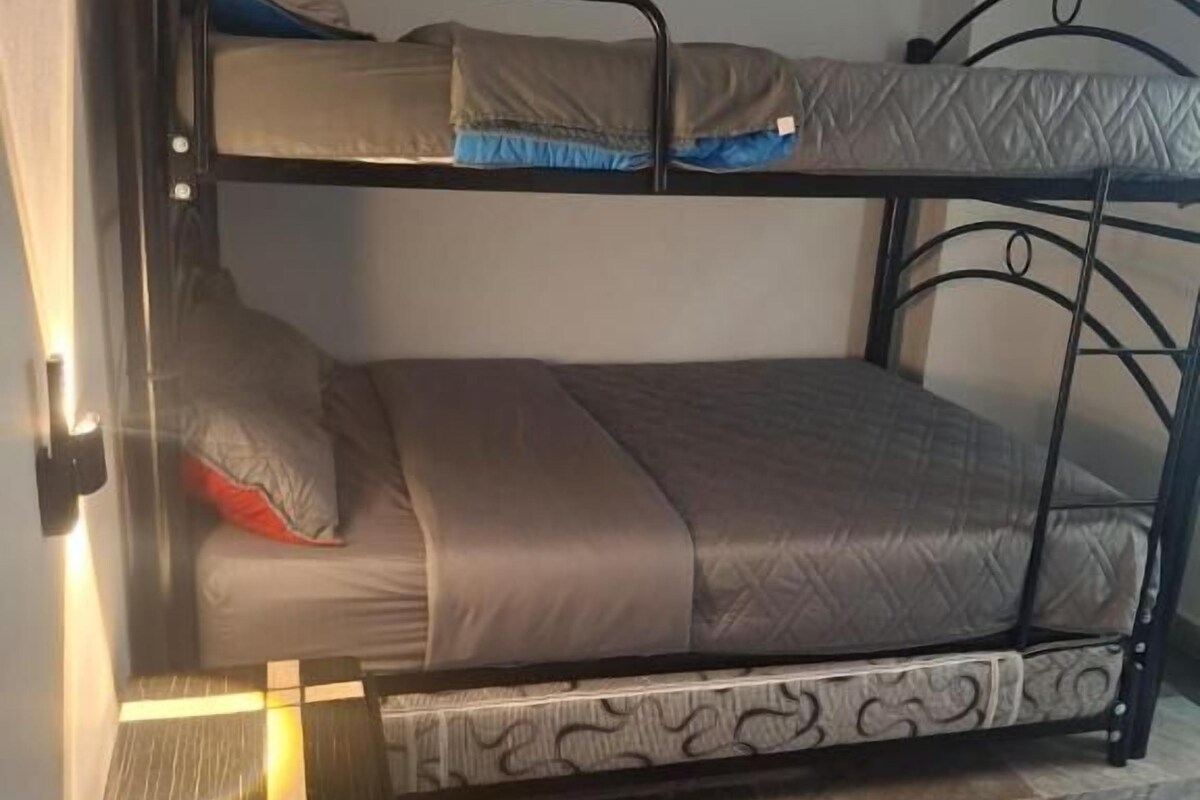 Een stapelbed met een matras en kussens, een nachtkastje met een lamp, en een getextureerd vloerkleed op een betegelde vloer.