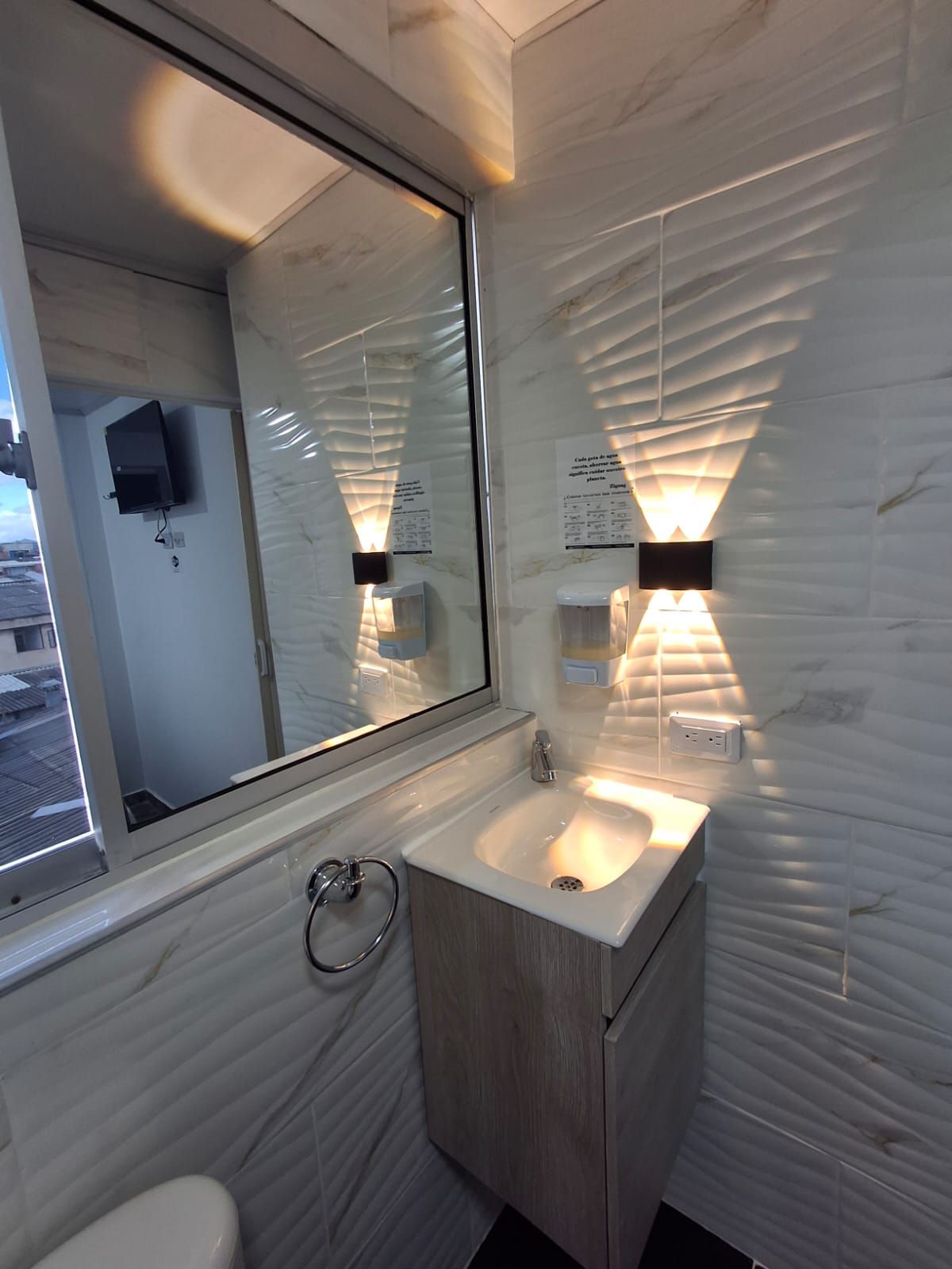 A bathroom with a large mirror, a sink, and a toilet. There are wall-mounted lights and a window.