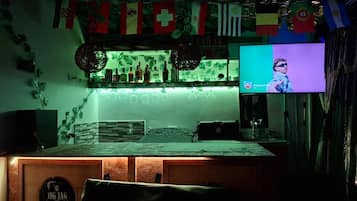 Un espace bar avec un comptoir, des étagères remplies de bouteilles et un écran de télévision diffusant une émission.