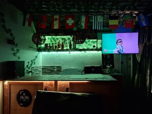 Un espace bar avec un comptoir, des étagères remplies de bouteilles et un écran de télévision diffusant une émission.