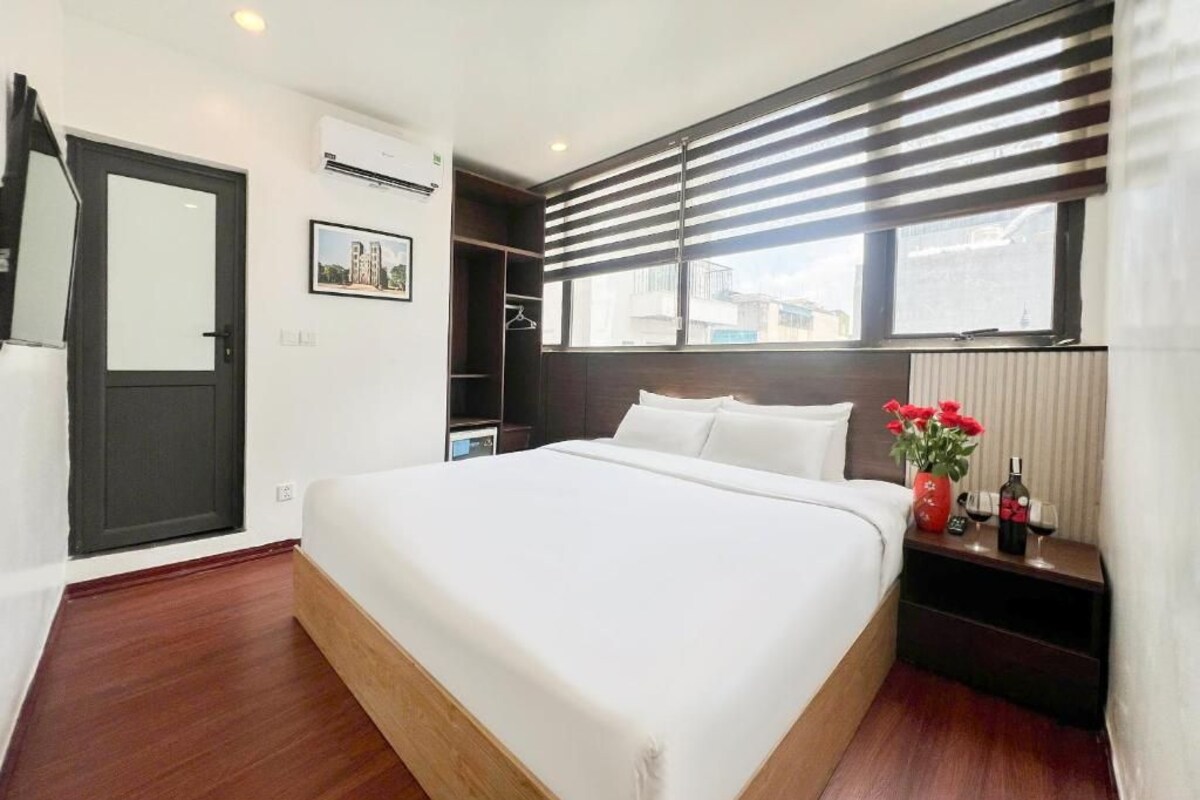 A hotel room with a large bed, a bedside table with a bottle and glasses, a window with blinds, and a door.