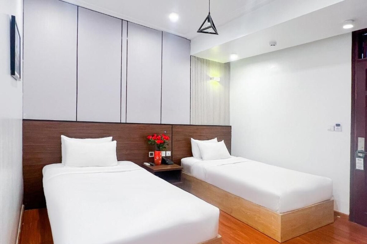A hotel room with two single beds, a wooden headboard, a nightstand, and a vase with red flowers.