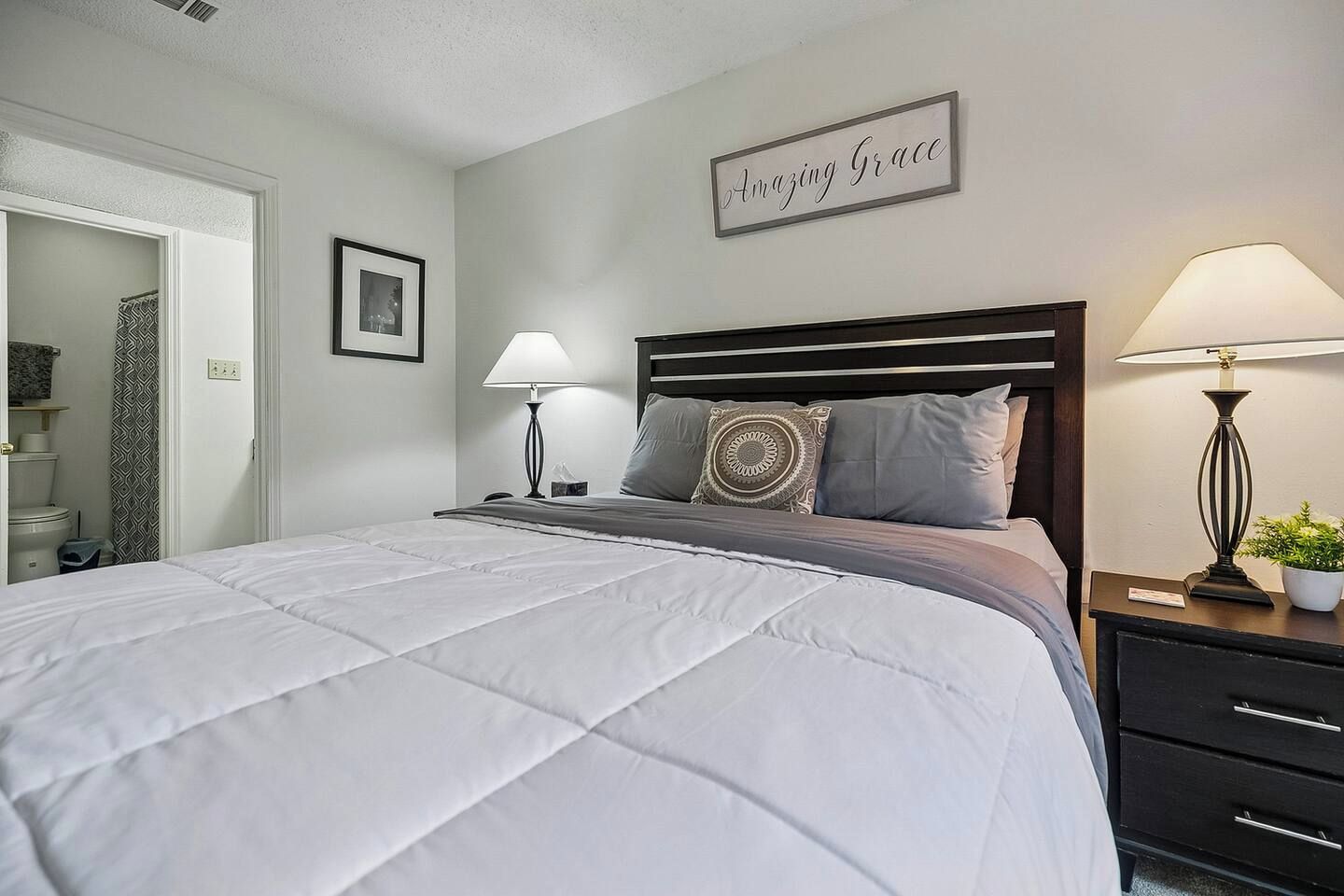 A neatly made bed with a headboard, two bedside lamps, a bedside table with a plant, and a wall sign.