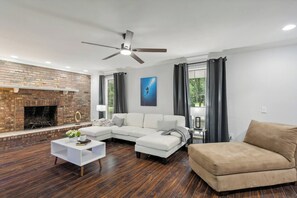 Ein modernes Wohnzimmer mit Kamin, Deckenventilator und einem großen Fenster mit Vorhängen.
