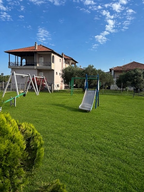 A grassy outdoor area with playground equipment, including swings and a slide, in front of a two-story building with a red-tiled roof. - Pancho Villa Family Apartments (Kallithea Pieras)