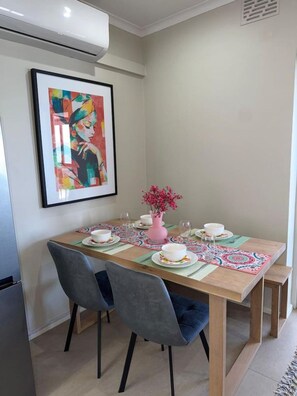 En matplats med ett bord dukat för två personer, ett inramat konstverk på väggen och en rosa vas med blommor.