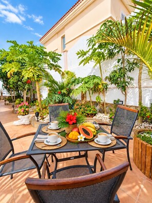 An outdoor patio with a table set for two, surrounded by tropical plants and a building with a tiled roof. - Villa Hills Meloneras (Maspalomas)