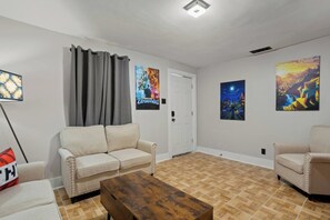 Ein Wohnzimmer mit einem beigen Sofa, einem Holztisch und zwei Sesseln. An der Wand hängen drei Poster, eines zeigt ein Minecraft-Spiel.