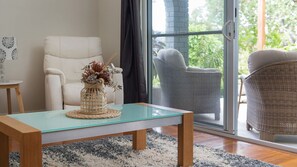 A modern living room with a glass-top coffee table, a white armchair, and a wicker chair with a view of greenery outside. - Grant Street Getaway (Port Macquarie)