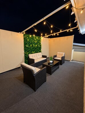 Una terraza en la azotea con mobiliario exterior, luces colgantes y un muro cubierto de plantas.