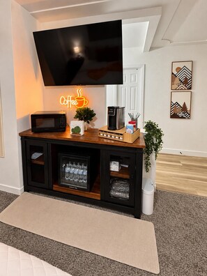 Sala de estar con televisor de pantalla plana en la pared, letrero de neón con temática de café, microondas, cafetera y un mueble con espacio de almacenamiento.