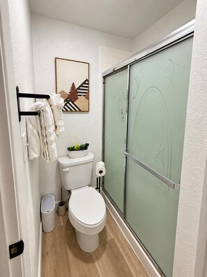 Un baño con inodoro, toallero con toallas, cubo de basura y cabina de ducha con mampara de cristal.