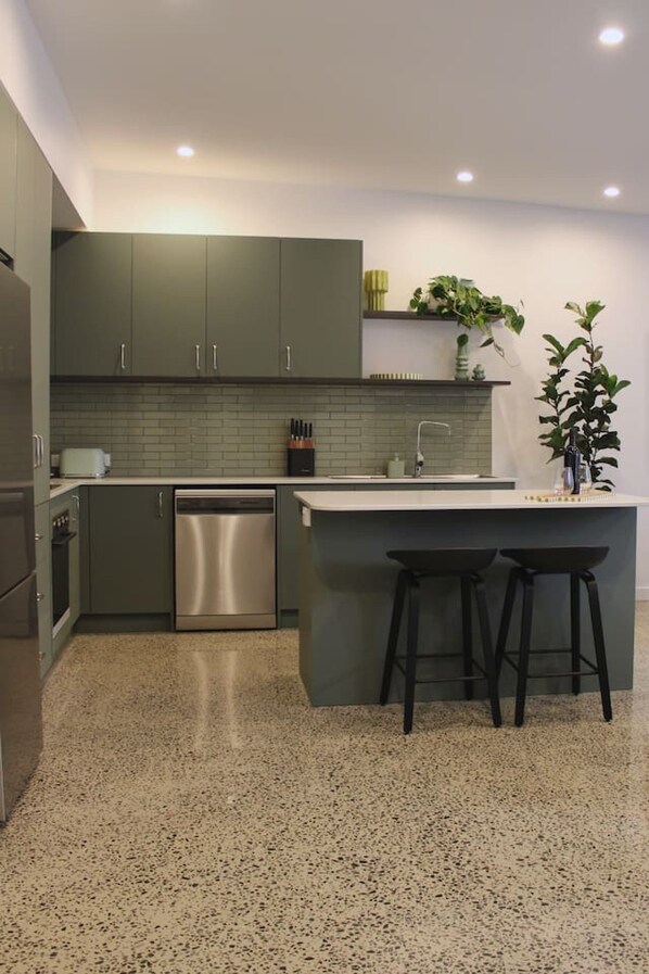A modern kitchen with grey cabinets, a stainless steel dishwasher, a built-in island with stools, and a green plant on the wall.