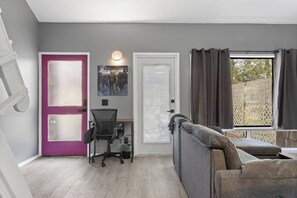 A room with a grey wall, a purple door, a white door, a black office chair, a grey sofa, a window with curtains, and a painting on the wall. - X Men Themed King Bed with Bidet (Gulfport)