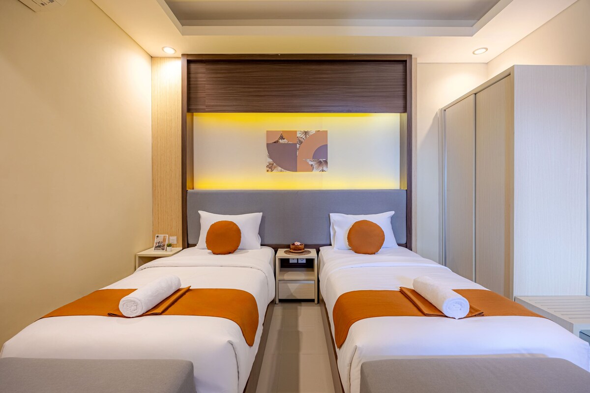 A hotel room with two single beds, a nightstand with a lamp, a small table with a bowl, and a wall-mounted artwork.