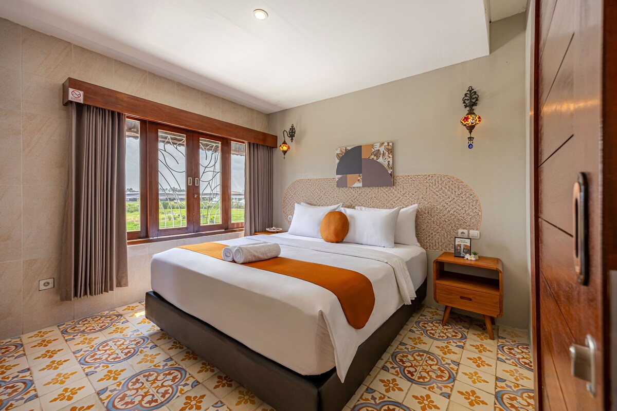 A hotel room with a large bed, a nightstand, a wall-mounted lamp, and a view of the outdoors through the window.