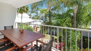 A balcony with a wooden table and chairs, surrounded by a white railing, overlooking a lush garden with various plants and palm trees.