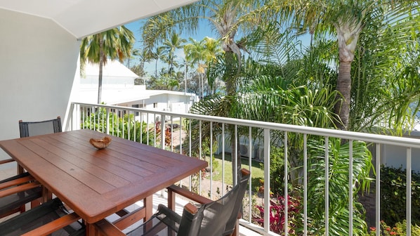 A balcony with a wooden table and chairs, surrounded by a white railing, overlooking a lush garden with various plants and palm trees.