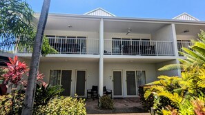 Un edificio de dos pisos con balcones, un área de estar al aire libre techada y una variedad de plantas tropicales.