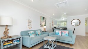 A modern living room with a sectional sofa, a small side table, a bookshelf, and a dining area with a kitchen in the background.