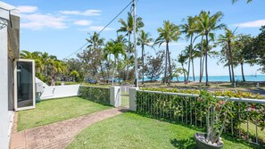 A residential property with a paved pathway leading to a gated entrance, surrounded by a well-maintained lawn and a variety of tropical plants.