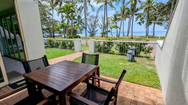 Un patio con una mesa y sillas de madera, un hidrante pequeño y vista al océano con palmeras.