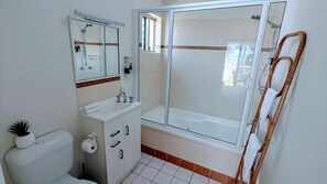 Un baño con ducha envuelta en vidrio, inodoro, lavamanos con espejo y una silla de madera.