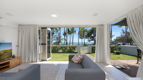 A modern living room with a sofa, a television, and a view of the ocean and palm trees.