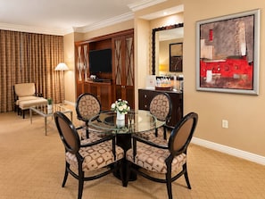 A hotel room with a dining area featuring a glass table and patterned chairs, a television in a wooden cabinet, and a painting on the wall. - MGM Signature 27F - F1 Track & Topgolf Views (Las Vegas)