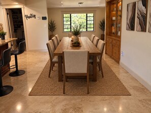Uma área de refeições com mesa e cadeiras de madeira, uma cozinha ao fundo e vista para o verde lá fora.