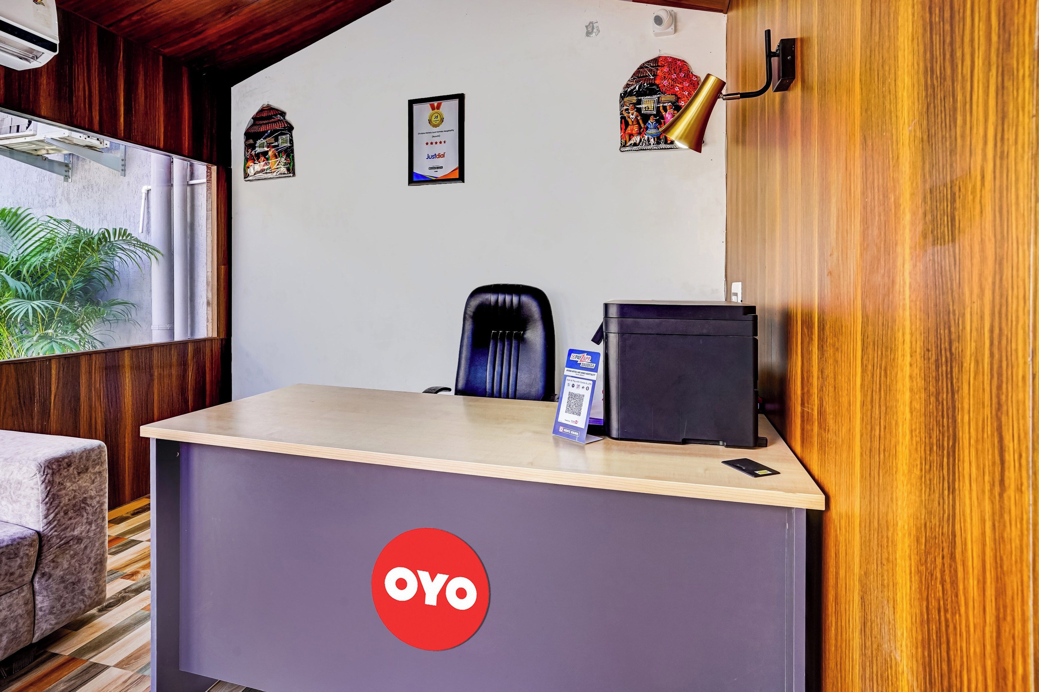 A reception desk with a 'OYO' logo, a framed certificate, a black office chair, a desk lamp, and a small table with a mobile phone.