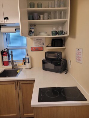 A kitchen with a built-in microwave, toaster, and induction cooktop. There is a shelf above the sink with various mugs and a fire extinguisher. - Quiet Studio · Free Parking (Hollywood)