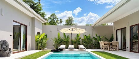 Uma área de piscina moderna, com piscina azul límpida, rodeada por vegetação exuberante e uma parede branca.
