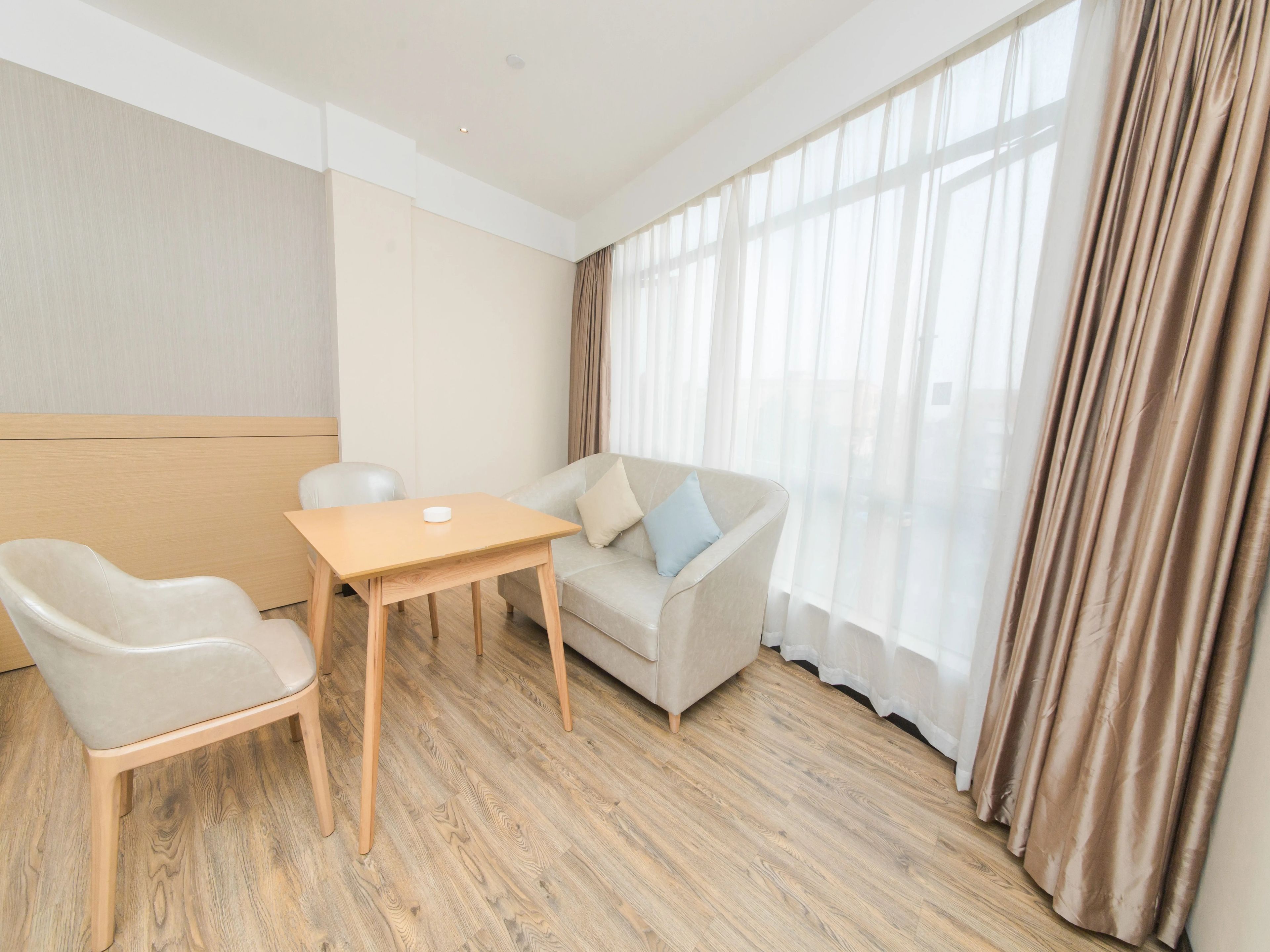 Una habitación de hotel moderna con suelo de madera, un sofá color beige, un sillón blanco y una pequeña mesa de madera.