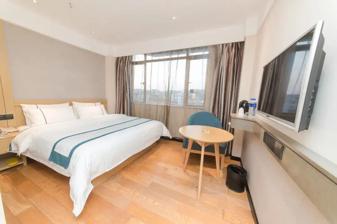 A hotel room with a bed, a small table, a chair, a TV, and a window with curtains.