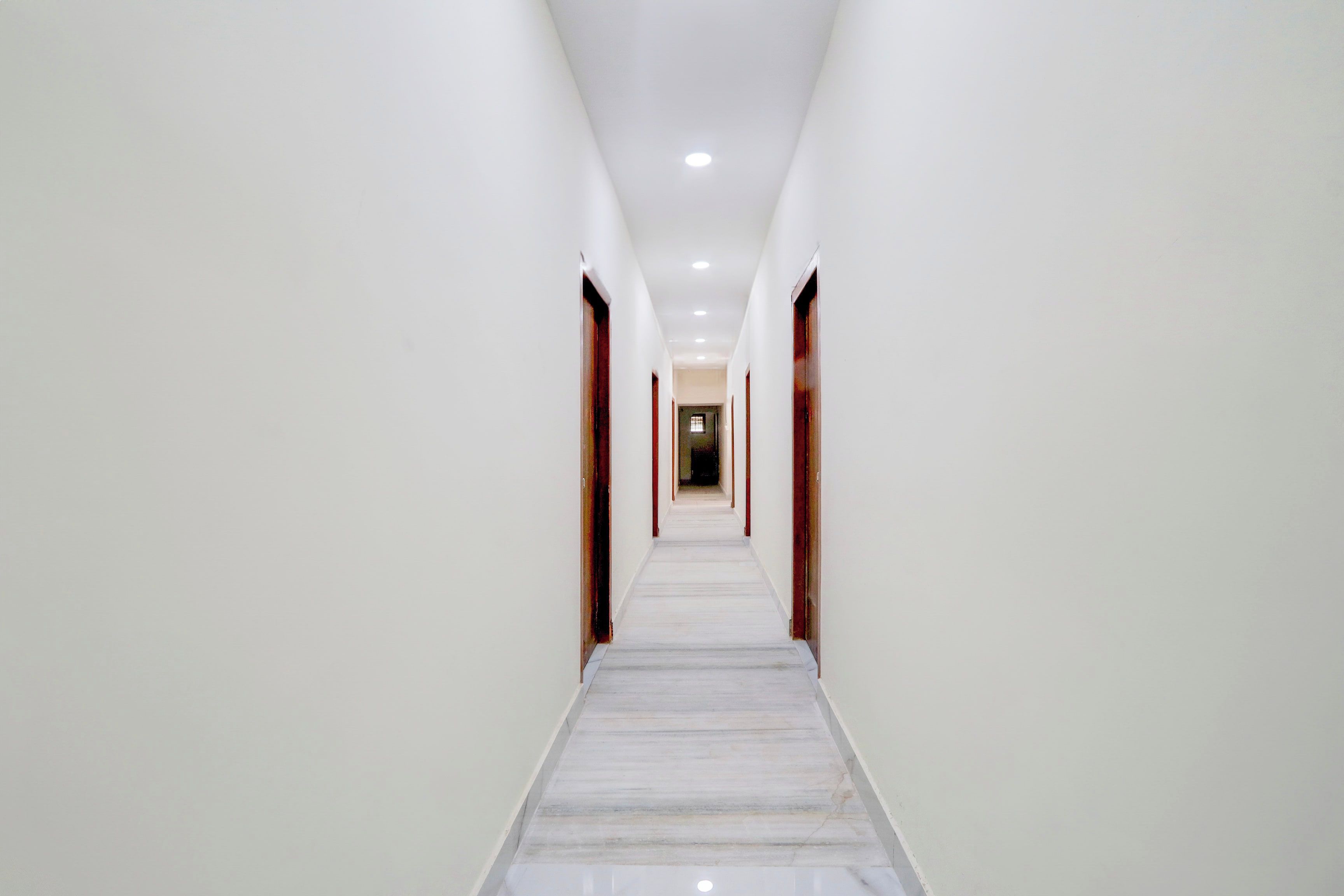 Un largo pasillo estrecho con paredes blancas y piso de mármol, que conduce a una puerta al final.