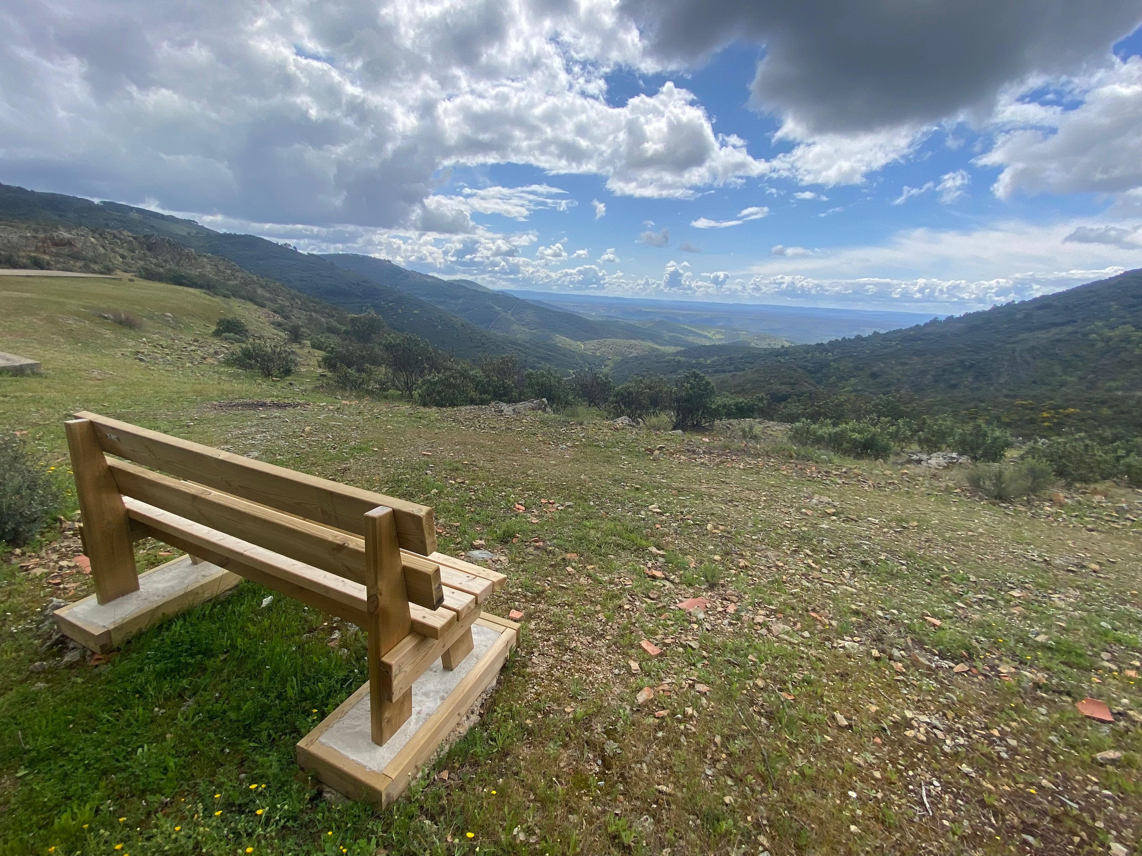 A wooden bench on a grassy hill with a view of distant hills and a partly cloudy sky.