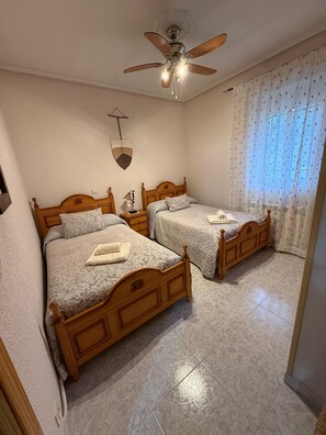 Un dormitorio con dos camas de madera, un ventilador de techo, un ventilador de pared y una ventana con cortinas traslúcidas.