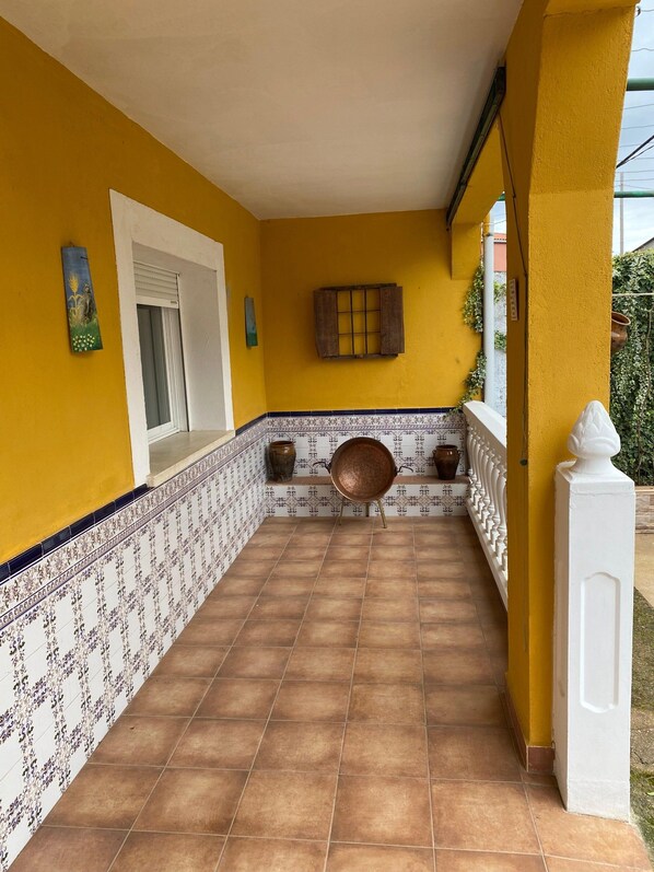Un espacio exterior techado con paredes amarillas, piso de baldosas y una silla metálica decorativa.