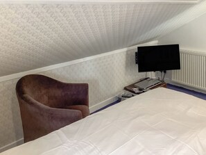 Ein Schlafzimmer mit einem Bett, einem braunen Sessel, einem Fernseher und einem kleinen Tisch mit elektronischen Geräten.