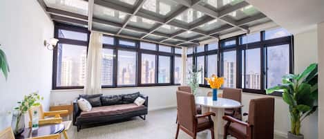 Una sala moderna con sofá, mesa de comedor, sillas y ventanas amplias con vista a la ciudad.