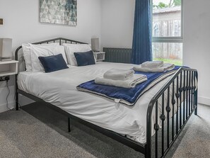 Ein ordentlich bezogenes Bett mit weißen Laken und blauen Kissen, einer blauen Tagesdecke und gefalteten Handtüchern auf einem Metallgestell in einem Zimmer mit weißen Wänden und einem Fenster mit blauen Vorhängen.
