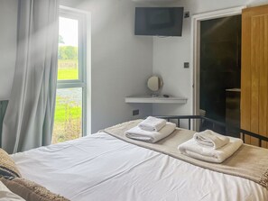 Quarto de hotel com cama, televisão fixada na parede, prateleira pequena com espelho e vista para o exterior pela janela.