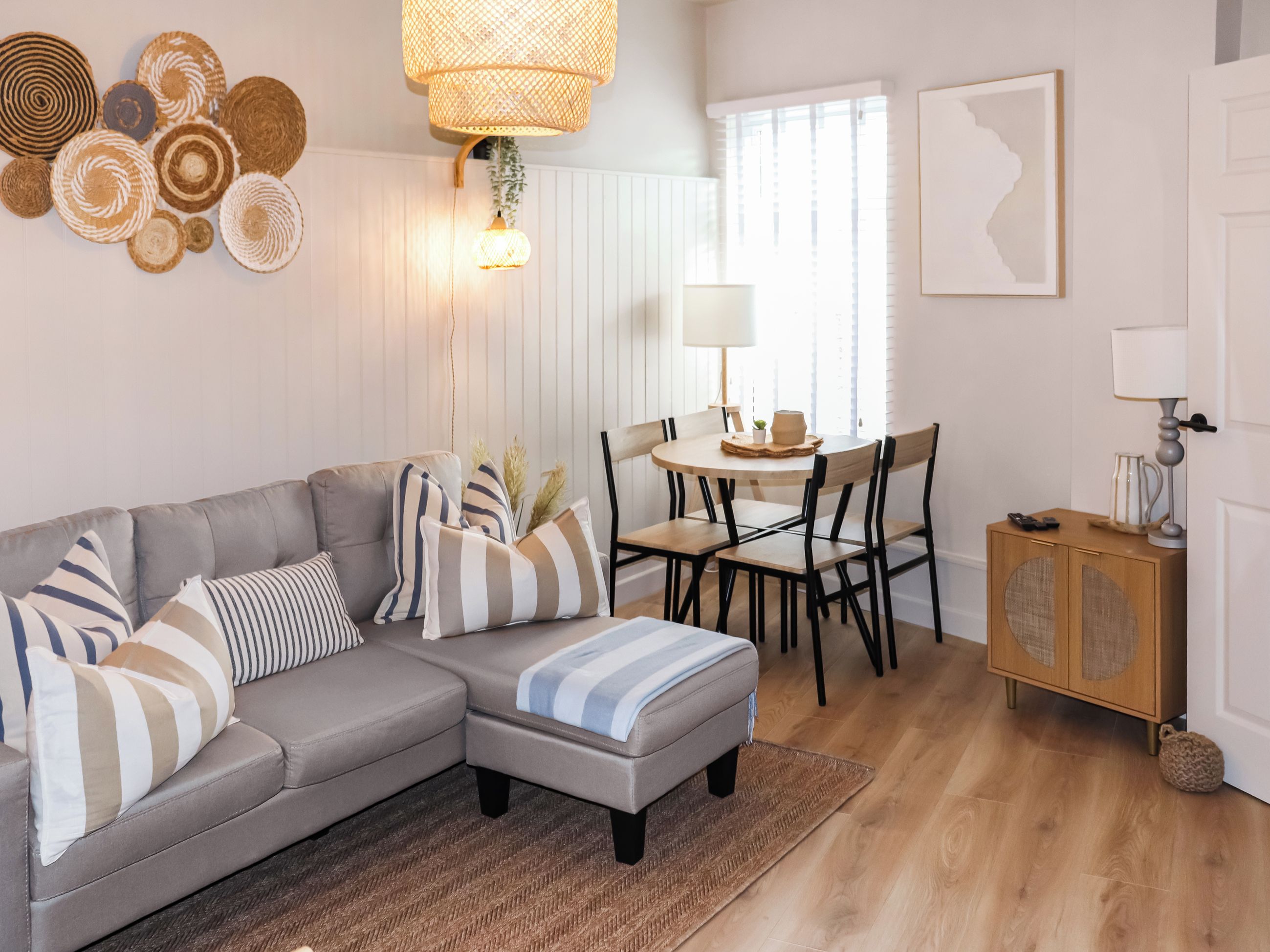 Ein Wohnzimmer mit grauem Ecksofa, einem runden Esstisch mit Stühlen, einer Holzkommode und einer Pendelleuchte.