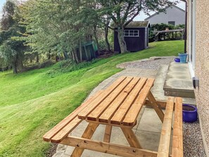 Une table de pique-nique en bois sur une terrasse en béton, avec une pelouse verdoyante et un petit abri de jardin en arrière-plan.