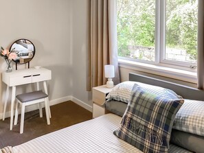 Une chambre à coucher avec un lit, une table de chevet avec une lampe, un petit bureau blanc avec un miroir et une fenêtre donnant sur un paysage verdoyant.