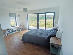 A bedroom with a large bed, a wooden floor, a bookshelf, and a view through a sliding door. - Welcome to Otter House a beautiful new build bungalow, located in Sand, Laide. (Laide)