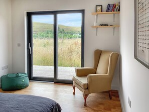 Ein Zimmer mit einer Schiebetür zum Garten, einem beigen Sessel, einem Bett mit dunkler Bettdecke und einem Regal mit Büchern und einem gerahmten Bild.